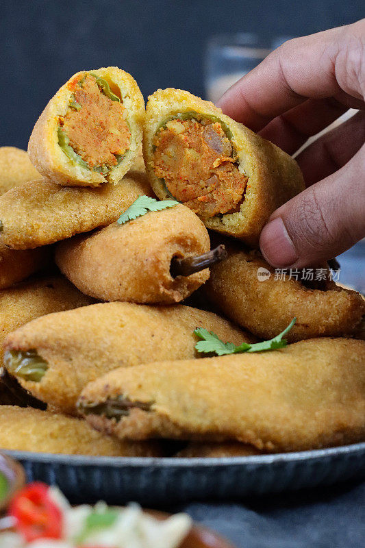 一个认不出来的人从盘子里挑出一堆绿辣椒馅饼(mirch pakora)的特写照片，炸辣椒里塞满了aloo mix(土豆泥、洋葱和马沙拉)，印度街头小吃，玻璃chai，灰色背景，前景焦点